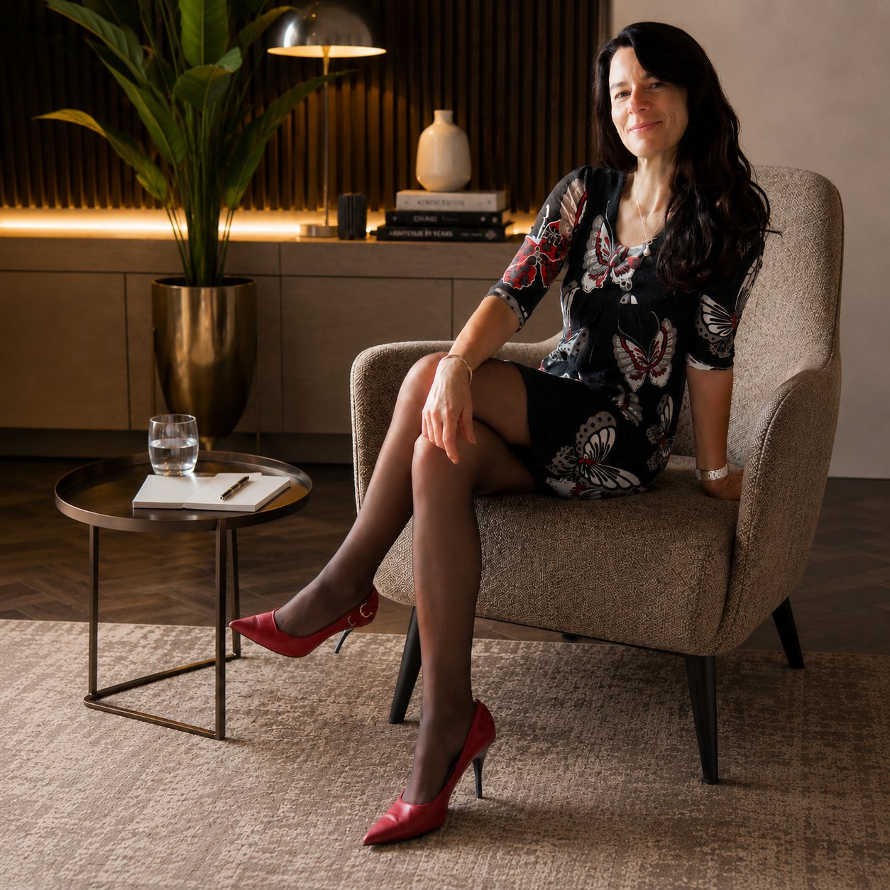 Amber Morrey seated in an armchair in a butterfly-print dress and red heels, in a warmly lit interior